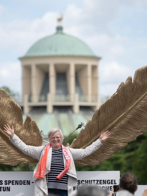 Eine mittelalte Frau schwingt lachend riesige goldene Flügel und stellt sich damit als einen "Schutzengel" dar.