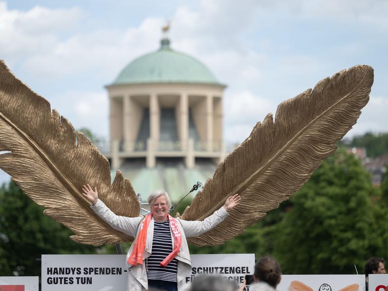 Eine mittelalte Frau schwingt lachend riesige goldene Flügel und stellt sich damit als einen "Schutzengel" dar.