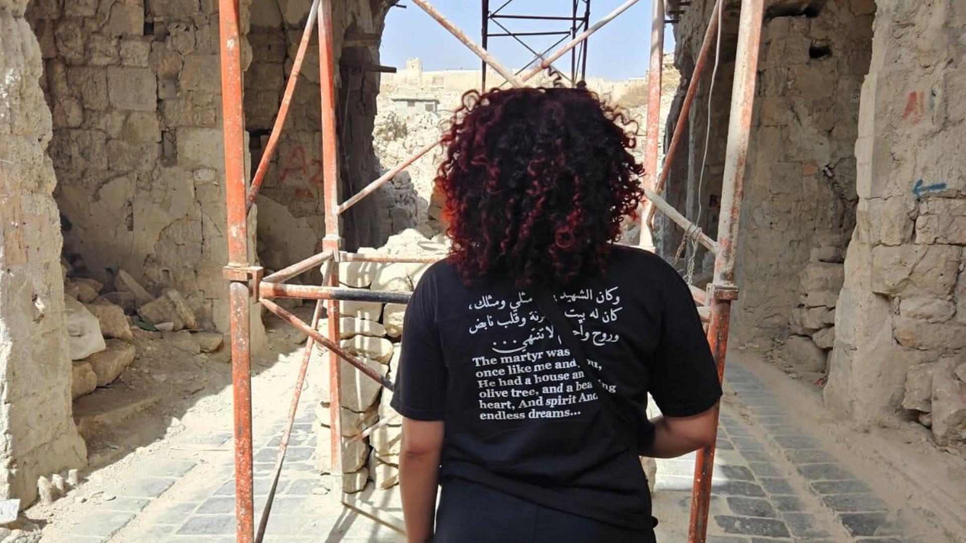 Hiba Obaid steht in der Altstadt von Aleppo. Sie dreht der Kamera den Rücken zu und trägt ein schwarzes Shirt mit einem aufgedruckten arabischen Text auf dem Rücken. Hiba Obaid steht in der Altstadt von Aleppo. Sie dreht der Kamera den Rücken zu und trägt ein schwarzes Shirt mit einem aufgedruckten arabischen Text auf dem Rücken.