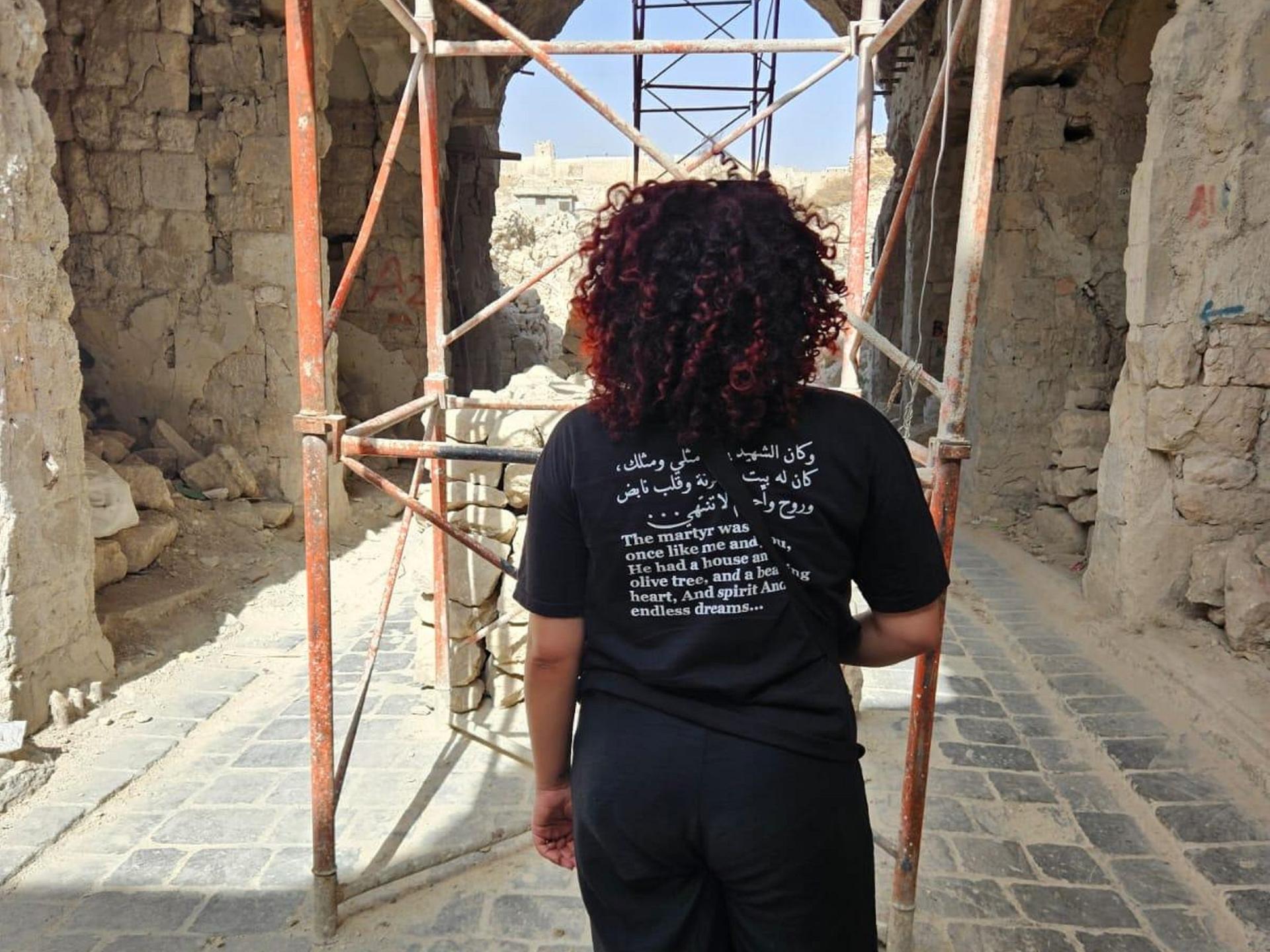 Hiba Obaid steht in der Altstadt von Aleppo. Sie dreht der Kamera den Rücken zu und trägt ein schwarzes Shirt mit einem aufgedruckten arabischen Text auf dem Rücken.