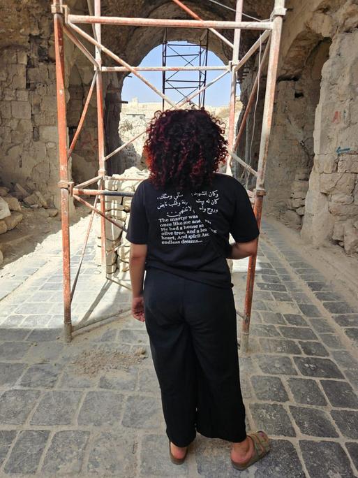 Hiba Obaid steht in der Altstadt von Aleppo. Sie dreht der Kamera den Rücken zu und trägt ein schwarzes Shirt mit einem aufgedruckten arabischen Text auf dem Rücken.
