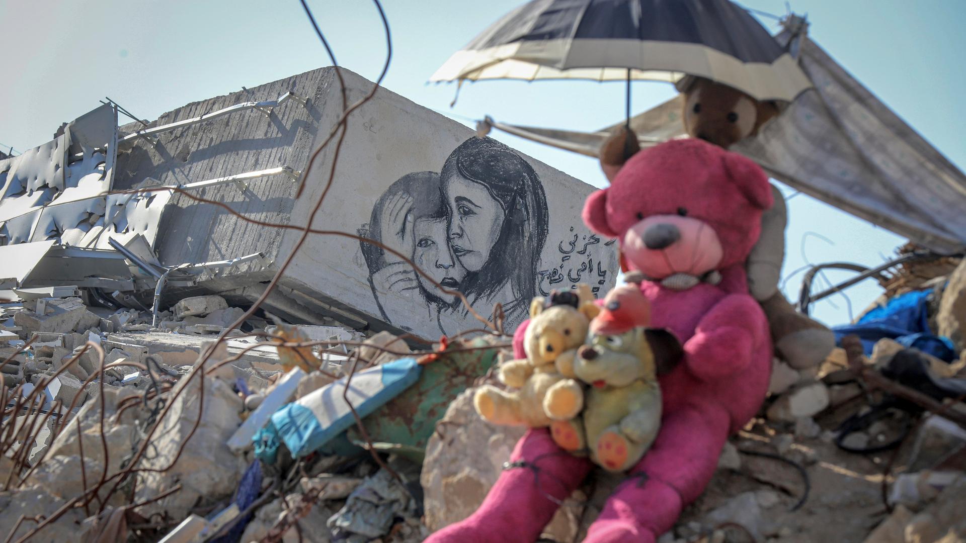 Teddybären sitzend auf einem Geröllhaufen eines eingestürzten Hauses in Gaza. Ein weiteres Stofftier hält einen kleinen Sonnenschirm über ihre Köpfe. Auf einem Stück Mauer ist eine Zeichnung auf der ein kleines Mädchen einen kleinen Jungen tröstend umarmt.