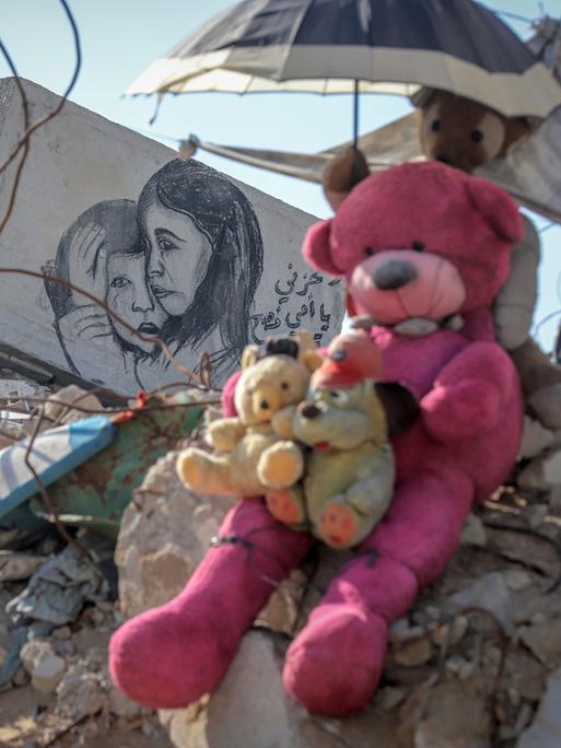 Teddybären sitzen auf einem Geröllhaufen eines eingestürzten Hauses in Gaza. Ein weiteres Stofftier hält einen kleinen Sonnenschirm über ihre Köpfe. Auf einem Stück Mauer ist eine Zeichnung auf der ein kleines Mädchen einen kleinen Jungen tröstend umarmt.