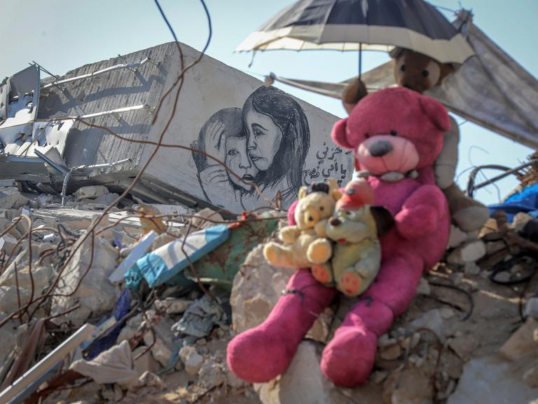 Teddybären sitzen auf einem Geröllhaufen eines eingestürzten Hauses in Gaza. Ein weiteres Stofftier hält einen kleinen Sonnenschirm über ihre Köpfe. Auf einem Stück Mauer ist eine Zeichnung auf der ein kleines Mädchen einen kleinen Jungen tröstend umarmt.