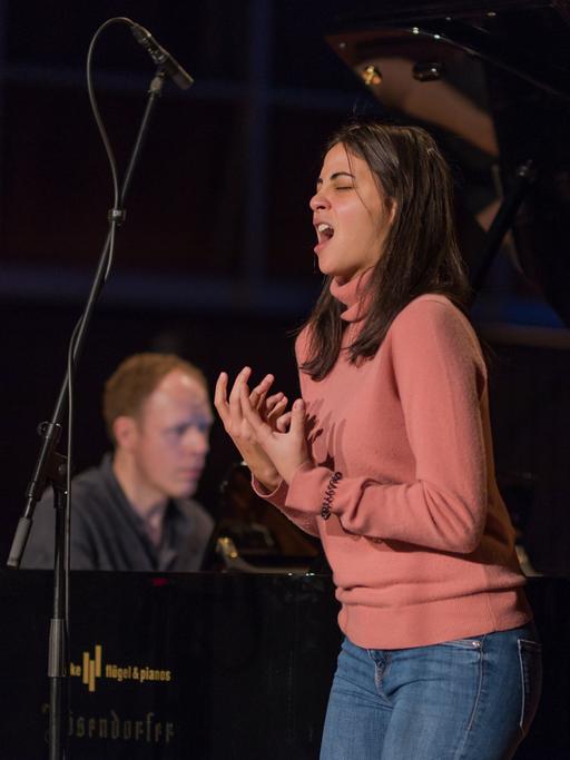 Eine junge Frau mit dunkelbraunen, schulterlangen Haaren singt ausdrucksstark. Sie formt mit den Händen eine Geste. Hinter ihr ist verschwommen der Pianist zu erkennen, der sie begleitet. Es sind die Sopranistin Fatma Said und der Pianist Joseph Middleton. 