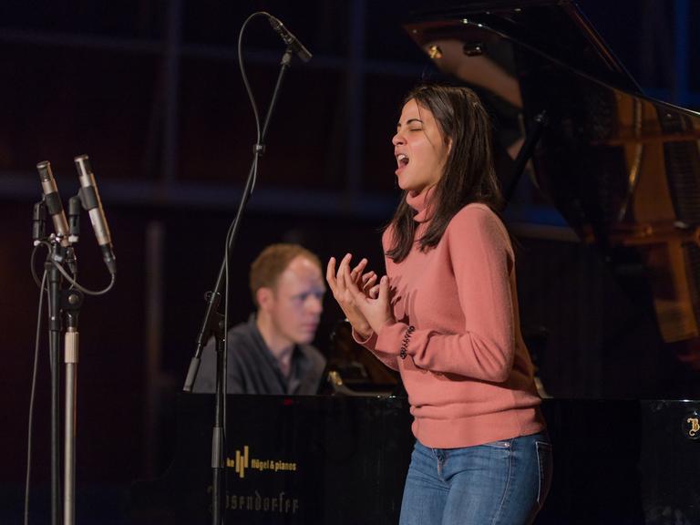 Eine junge Frau mit dunkelbraunen, schulterlangen Haaren singt ausdrucksstark. Sie formt mit den Händen eine Geste. Hinter ihr ist verschwommen der Pianist zu erkennen, der sie begleitet. Es sind die Sopranistin Fatma Said und der Pianist Joseph Middleton. 