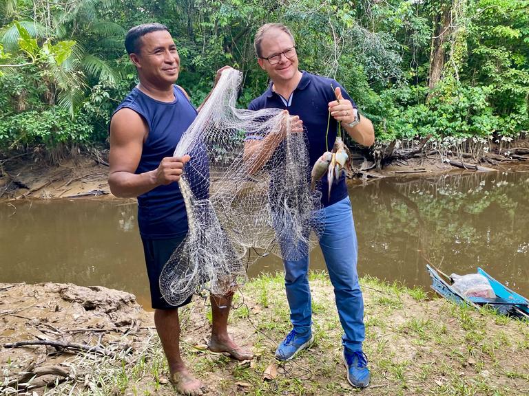 Bundesumweltminister Carsten Schneider (r, SPD) lässt sich von einem Bewohner der Quilombola-Gemeinschaft Abacatal Fische zeigen, die der örtliche Lehrer eben mit einem Netz im Fluss gefangen hat.