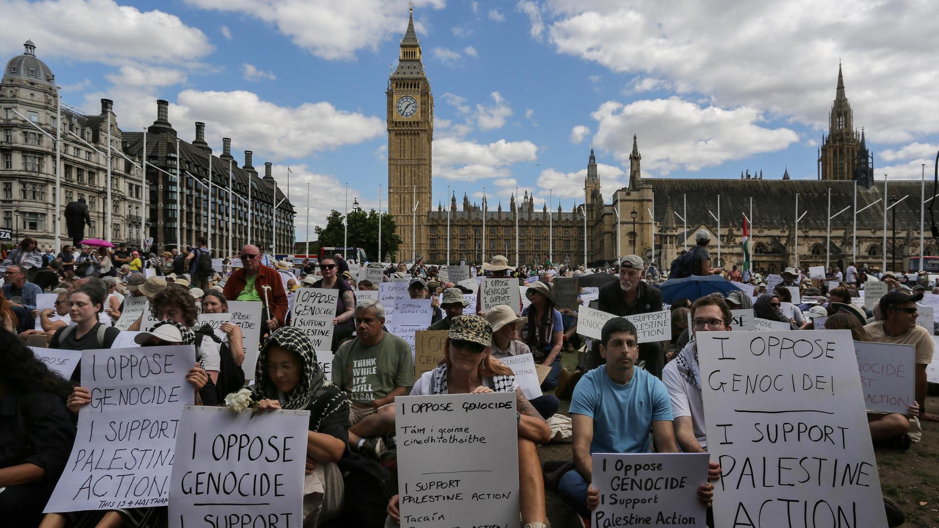 Menschen sitzen vor dem britischen Parlament in London, Big Ben ist zu sehen. Auf ihren Plakaten steht: "I oppose genozide, I support Palestine Action"