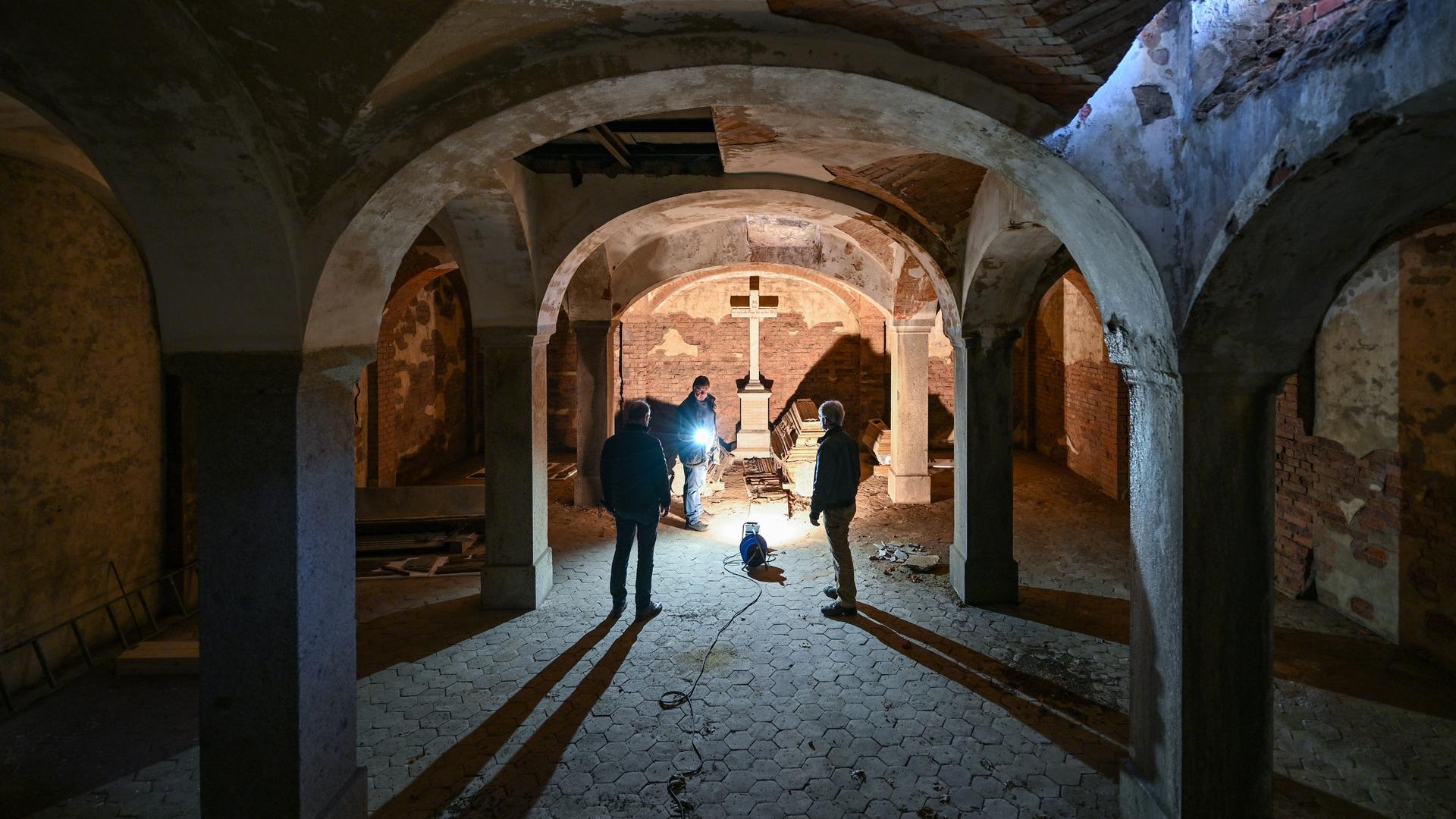 Blick in eine Gruft mit Tonnengewölbe. Drei Personen stehen in einer dunklen, nur punktuell beleuchteten Gruft, in deren Mitte ein Kreuz steht.