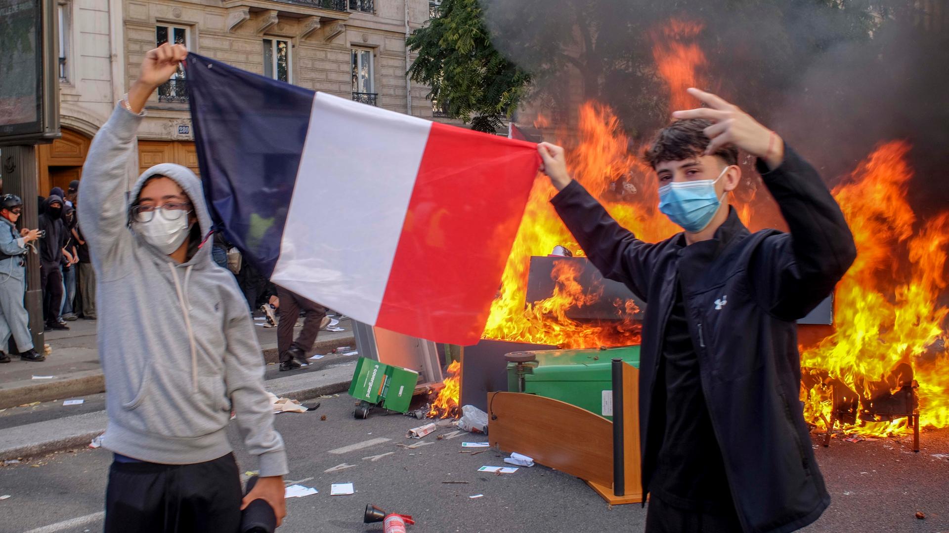 Zwei Schüler halten eine französiche Flagge, sie tragen Atemmasken und hinter ihnen auf der Straße brennt eine Barrikade aus umgestürzten Tischen, Stühlen und Mülltonnen.