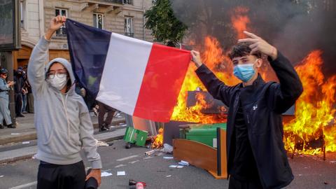 Zwei Schüler halten eine französiche Flagge, sie tragen Atemmasken und hinter ihnen auf der Straße brennt eine Barrikade aus umgestürzten Tischen, Stühlen und Mülltonnen.