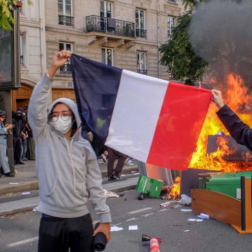 Zwei Schüler halten eine französische Flagge, sie tragen Atemmasken und hinter ihnen auf der Straße brennt eine Barrikade aus umgestürzten Tischen, Stühlen und Mülltonnen.