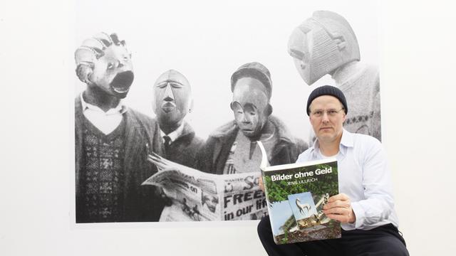 Der Künstler Jens Ullrich steht vor einem großformatigen Foto, das Menschen mit Masken zeigt