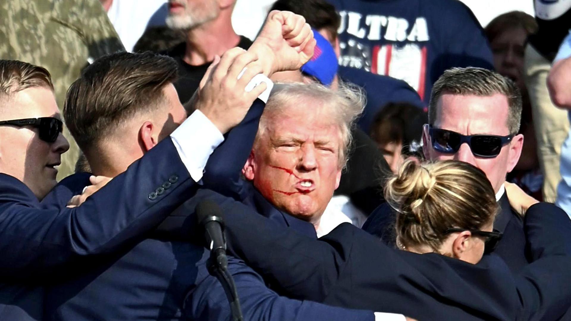 Nach einem Attentat auf ihn reckt Donald Trump die Faust, im Gesicht hat er Blutspritzer. Er wird von Secret-Service-Agenten umringt.