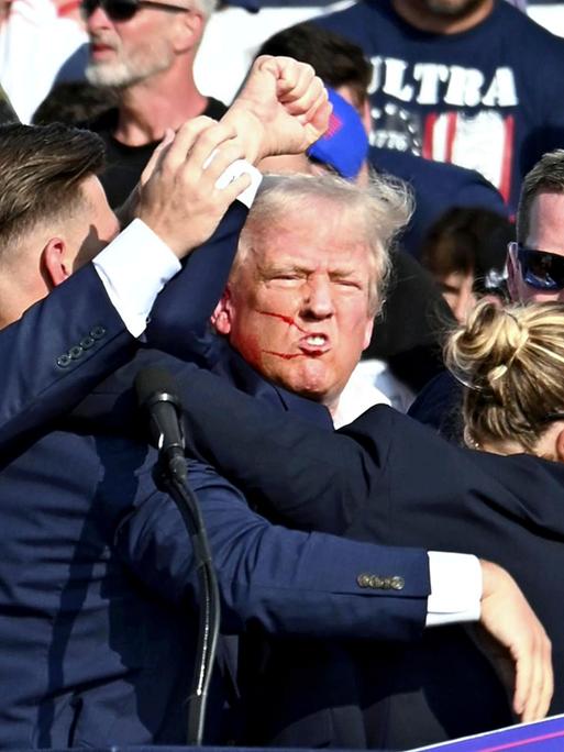Nach einem Attentat auf ihn reckt Donald Trump die Faust, im Gesicht hat er Blutspritzer. Er wird von Secret-Service-Agenten umringt.