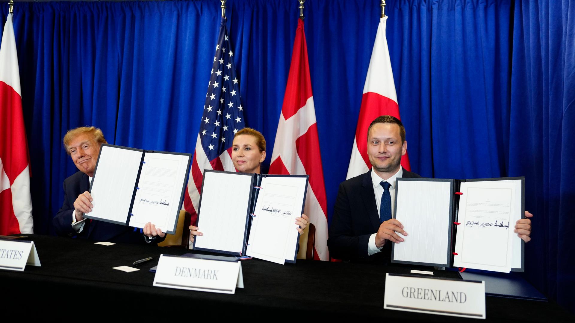 Nach der Unterzeichnung des Sicherheitsabkommens - US-Präsident Trump (l.), Dänemarks Ministerpräsidentin Frederiksen und Grönlands Regierungschef Nielsen; sie sitzen an einem langen Tisch und halten jeweils ein Dokument in die Höhe. Nach der Unterzeichnung des Sicherheitsabkommens - US-Präsident Trump (l.), Dänemarks Ministerpräsidentin Frederiksen und Grönlands Regierungschef Nielsen; sie sitzen an einem langen Tisch und halten jeweils ein Dokument in die Höhe.
