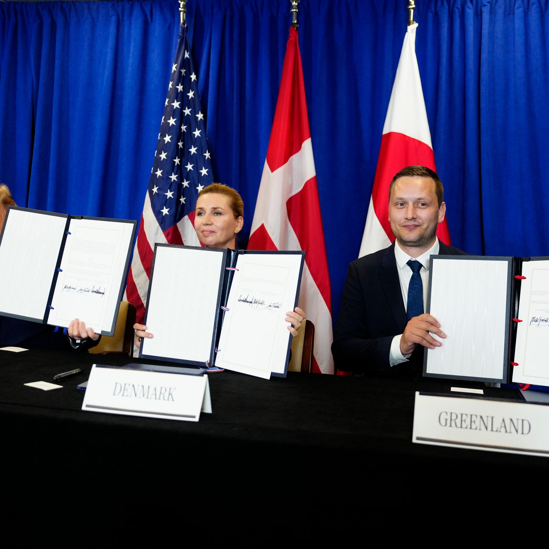 Nach der Unterzeichnung des Sicherheitsabkommens - US-Präsident Trump (l.), Dänemarks Ministerpräsidentin Frederiksen und Grönlands Regierungschef Nielsen; sie sitzen an einem langen Tisch und halten jeweils ein Dokument in die Höhe.