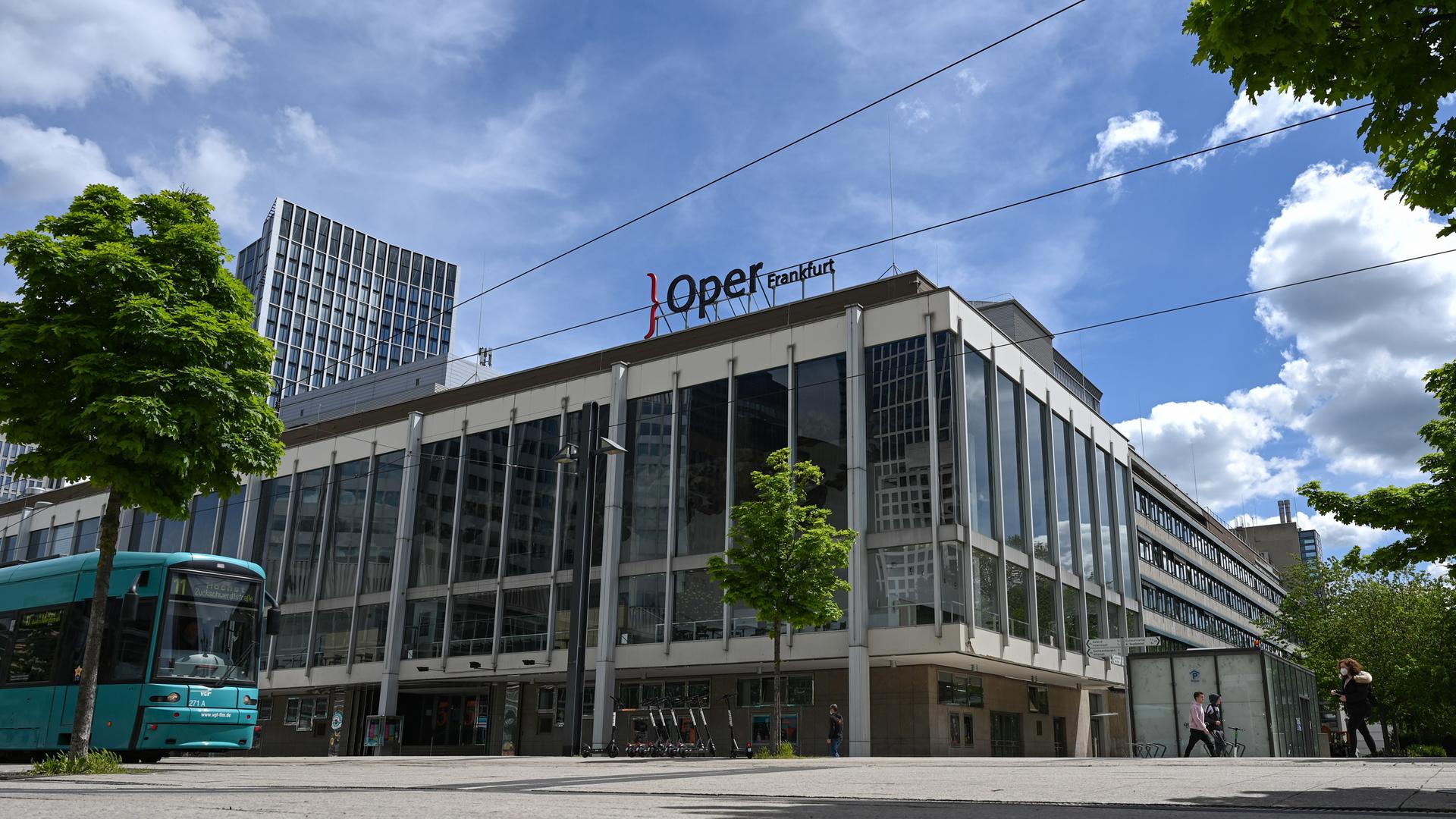 Das Gebäude der Oper und des Schauspielhauses Frankfurt am Main. Auf dem Dach ist der Schriftzug "Oper Frankfurt" zu sehen. Am linken Bildrand ist eine Straßenbahn zu sehen.