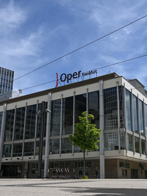 Das Gebäude der Oper und des Schauspielhauses Frankfurt am Main. Auf dem Dach ist der Schriftzug "Oper Frankfurt" zu sehen. Am linken Bildrand ist eine Straßenbahn zu sehen.