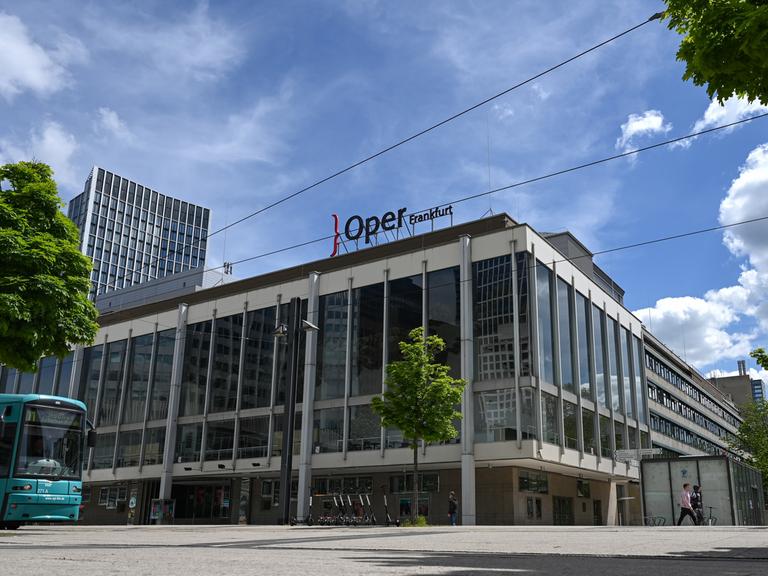 Das Gebäude der Oper und des Schauspielhauses Frankfurt am Main. Auf dem Dach ist der Schriftzug "Oper Frankfurt" zu sehen. Am linken Bildrand ist eine Straßenbahn zu sehen.