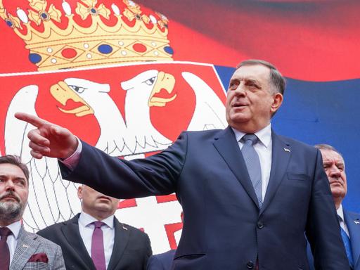 Der bosnisch-serbische Präsident Milorad Dodik steht vor einer Flagge der Republika Srpska in einem schwarzen Sakko. Mit seiner rechten Hand zeigt er links aus dem Bild heraus. Sein Blickist ernst.