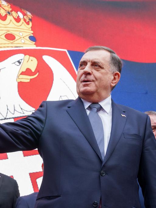 Der bosnisch-serbische Präsident Milorad Dodik steht vor einer Flagge der Republika Srpska in einem schwarzen Sakko. Mit seiner rechten Hand zeigt er links aus dem Bild heraus. Sein Blickist ernst.