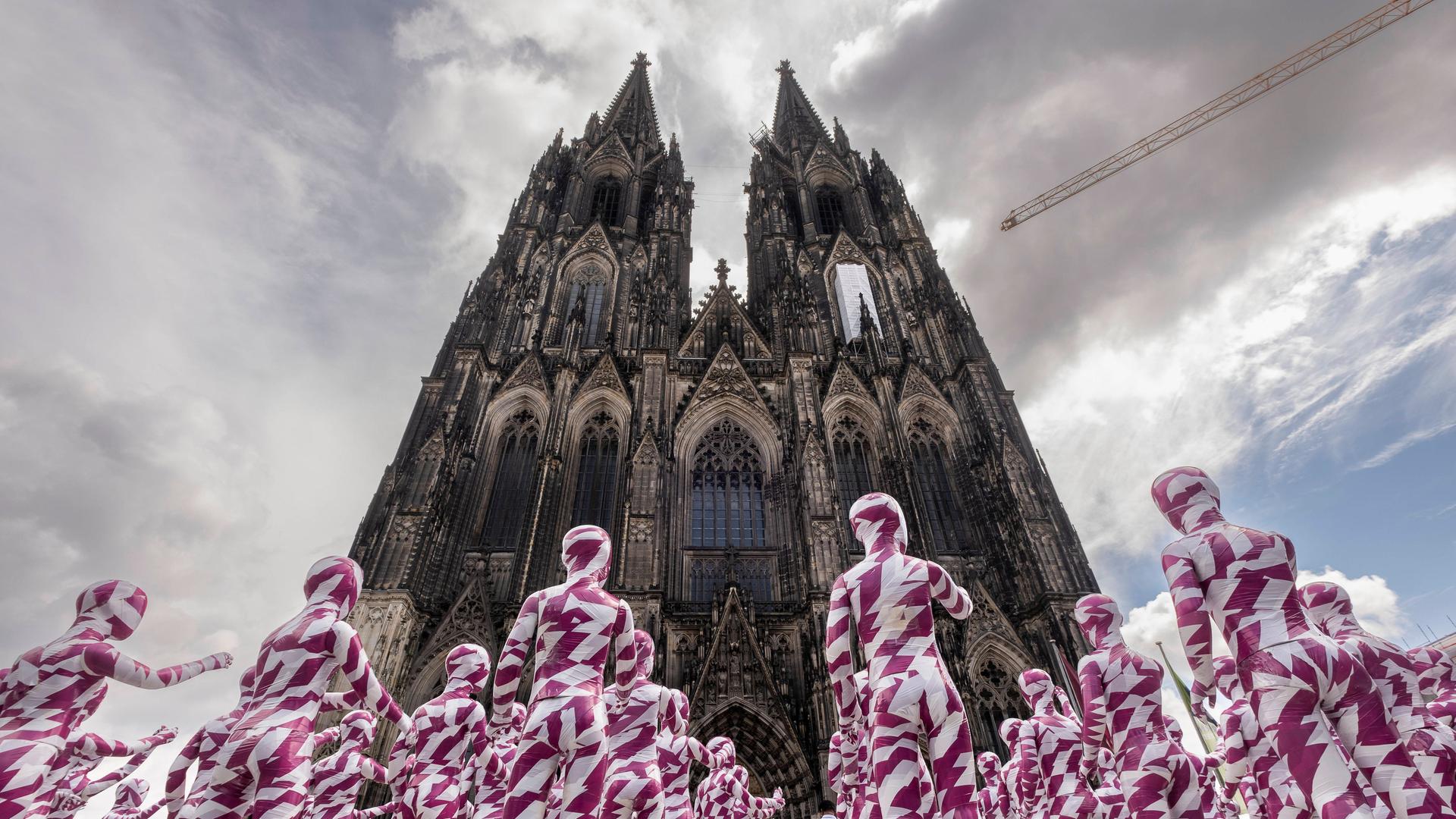333 kleine, in weiße und purpurfarbenene Bänder eingewickelte Schaufensterpuppen aufgestellt stehen vor dem Kölner Dom und weisen mit dem Finger auf ihn.