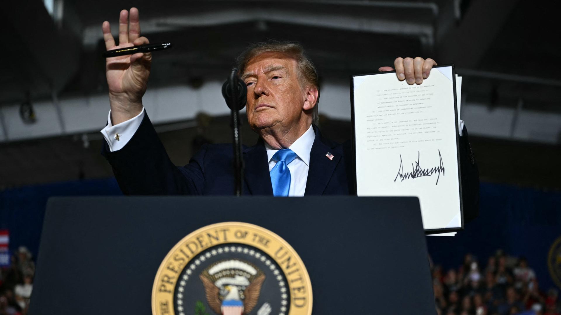 Donald Trump steht auf der Bühne vor einem Rednerpult. Er hält das unterzeichnete Dekret in die Höhe.