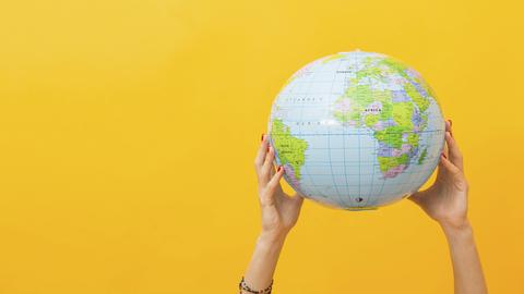 Zwei Hände halten Globus vor einem gelben Hintergrund in die Höhe.