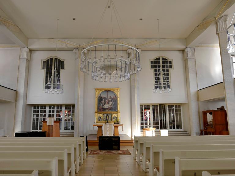 Das Schiff der Luisenkirche in Berlin. Bänke sind angereiht. Alles ist sehr hell. Hinter dem Altar ist ein goldgeschmücktes Bild.