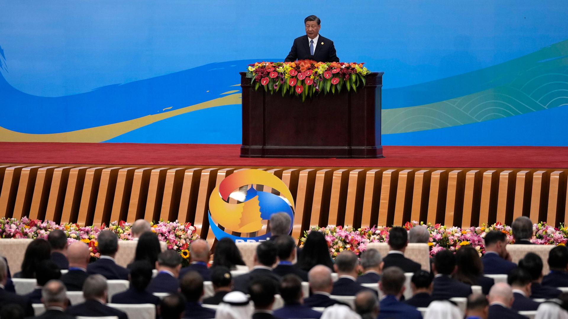 Der chinesische Präsident Xi Jinping steht hinter einem mit Blumen geschmückten Rednerpult. Im Vordergrund sitzen Teilnehmer und hören zu.
