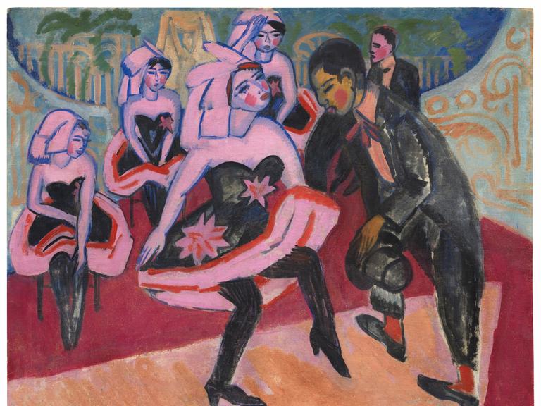 Das Bild zeigt das Gemälde "Tanz im Varieté" von Ernst Ludwig Kircher (1911). Es stellt eine Frau im kurzen Kleid dar, die mit einem Mann im Frack tanzt. Im Hintergrund sind ähnlich gekleidete Frauen zu sehen. Das Bild im expressionistischen Stil ist in bunten Farben gemalt. 