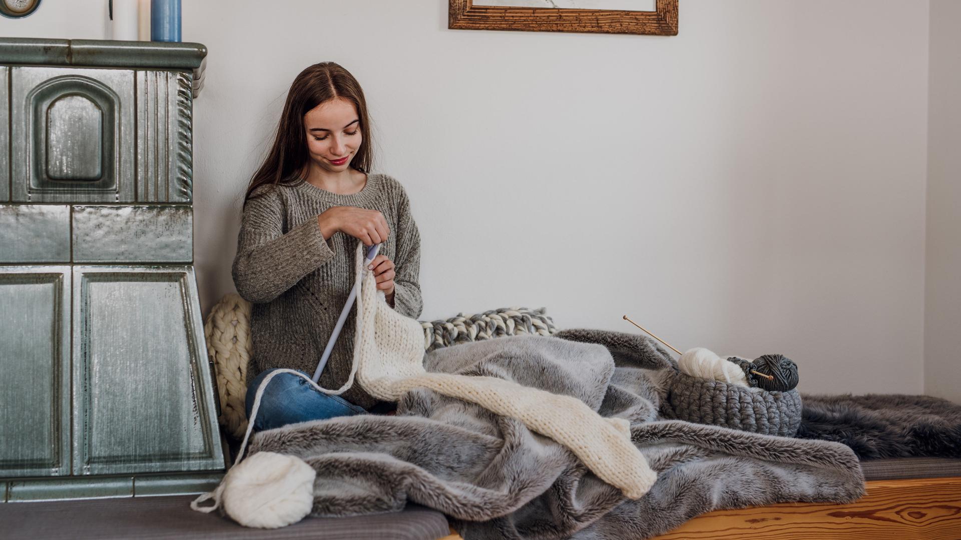 Eine junge Frau sitzt am Kachelofen und strickt.