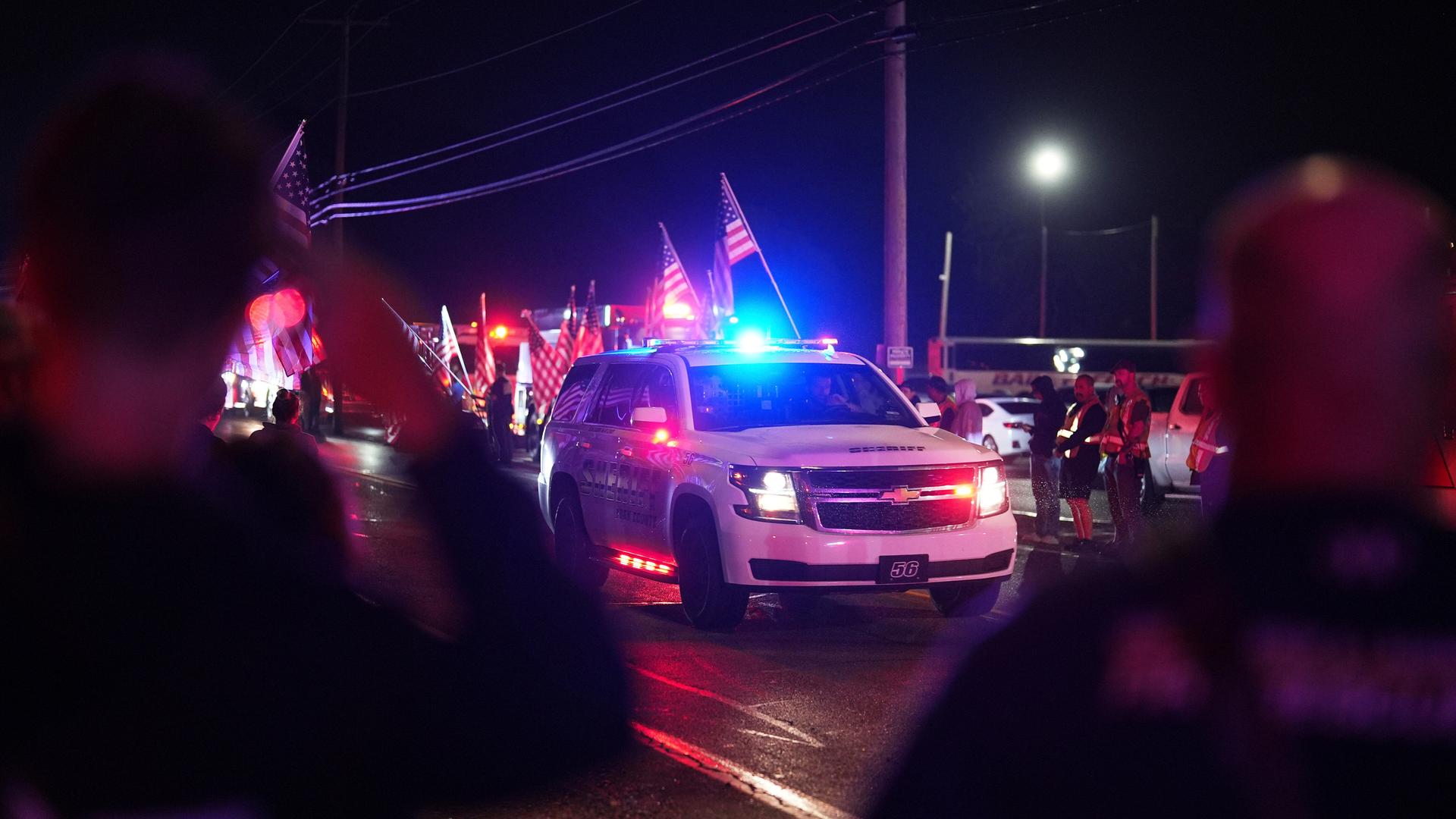 Ein Sheriff-Auto mit eingeschaltetem Blaulicht fährt bei Dunkelheit an Menschen vorbei, die USA-Flaggen halten.