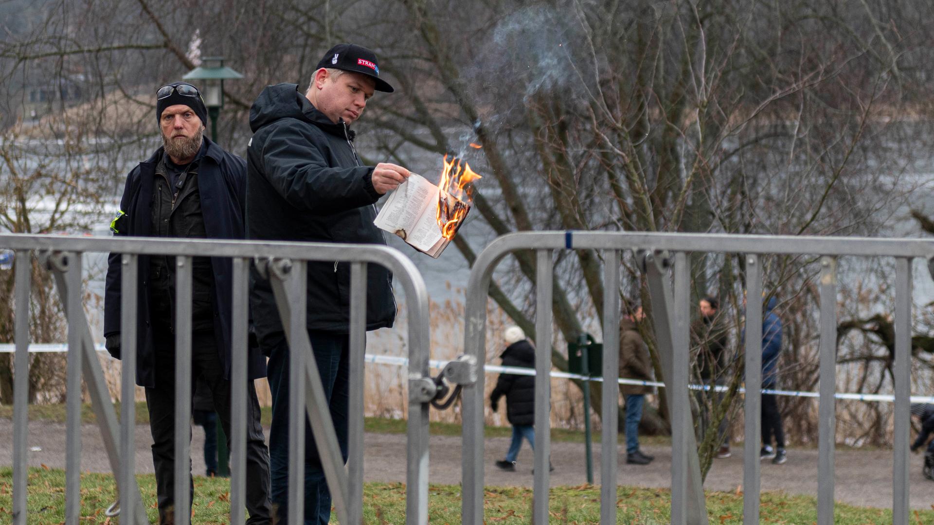 Paludan zündet auf einer Wiese nahe eines Flusses gerade Seiten des Koran an. Davor zwei Absperrgitter. Im Hintergrund sind weitere Menschen zu sehen. Paludan zündet auf einer Wiese nahe eines Flusses gerade Seiten des Koran an. Davor zwei Absperrgitter. Im Hintergrund sind weitere Menschen zu sehen.