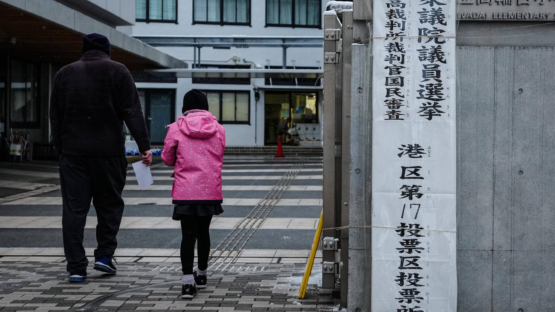 Ein dunkel gekleideter Mann und eine Frau in einem pinken Anorak gehen in Tokio in ein Wahllokal. Ein dunkel gekleideter Mann und eine Frau in einem pinken Anorak gehen in Tokio in ein Wahllokal.
