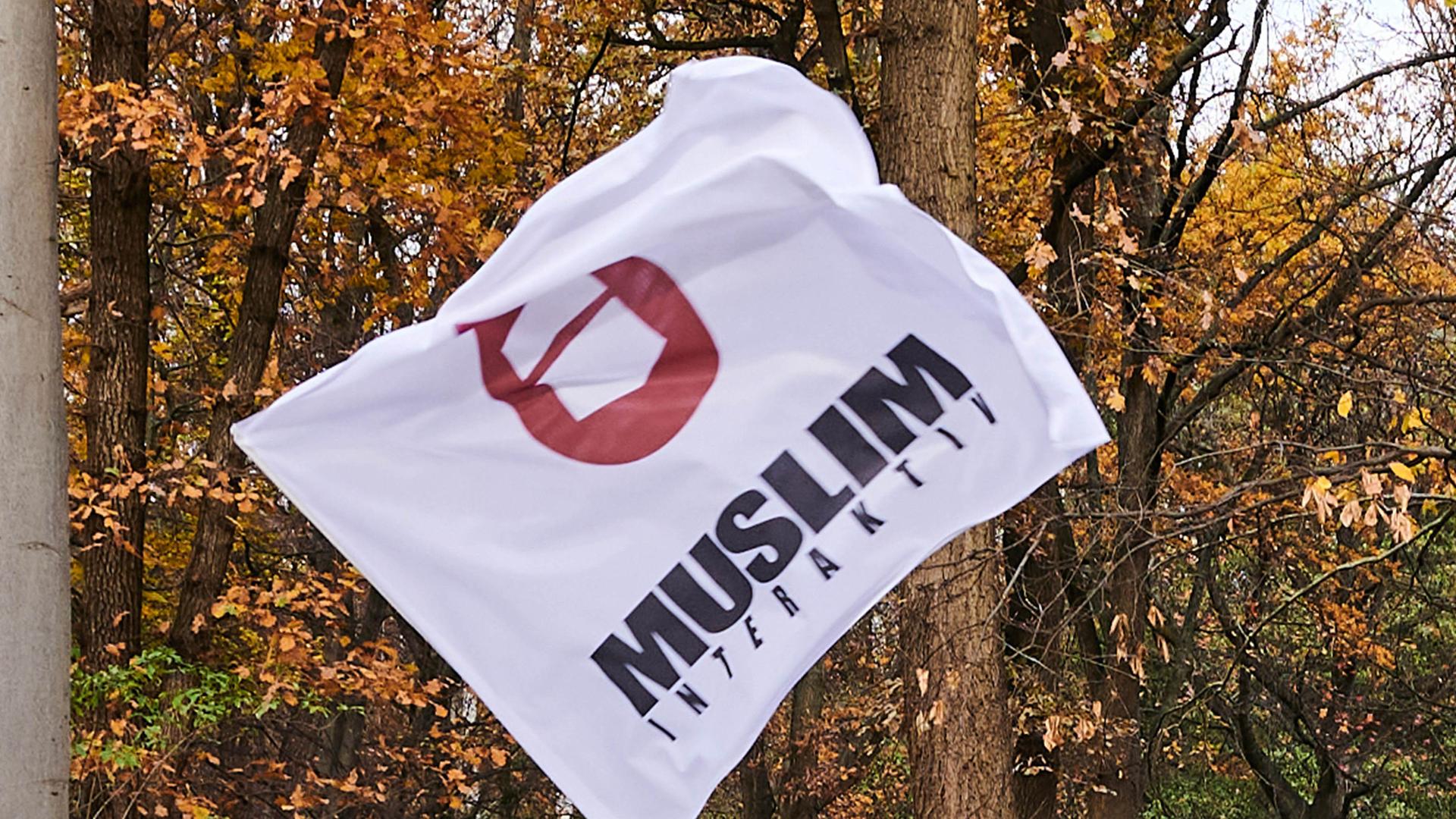 "Muslim Interaktiv" steht auf einer Fahne. Im Hintergrund sind Bäume, deren Blätter herbstlich gelb-braun sind. 