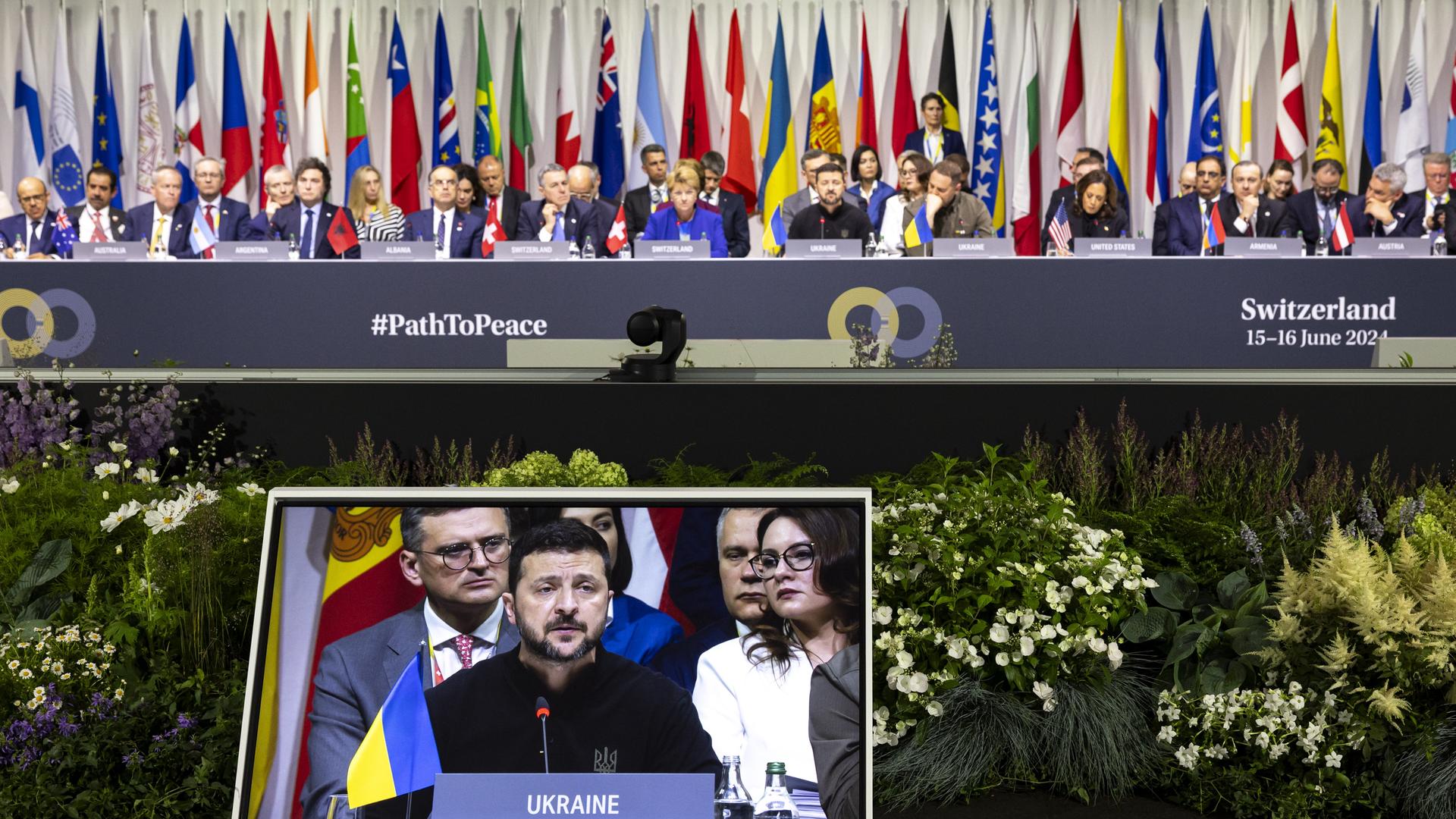 Wolodymyr Selenskyj, Präsident der Ukraine, spricht während einer Friedenskonferenz in der Schweiz. Er ist auf einem Bildschirm zu sehen, hinter ihm sind Teilnehmer und Flaggen aus zahlreichen Ländern zu erkennen. 