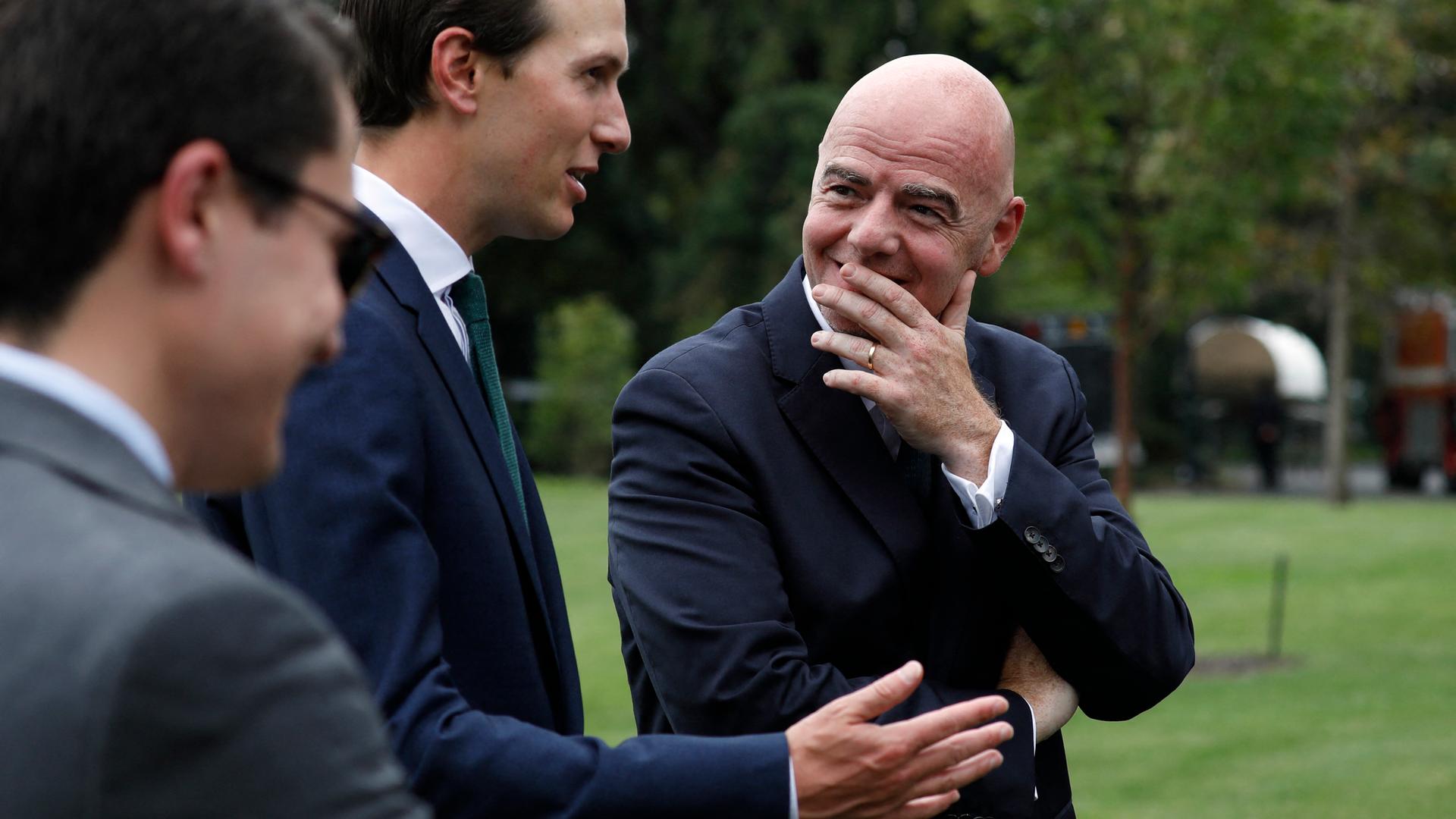 Zwei Männer in Anzügen unterhalten sich. Sie stehen auf einem Rasenstück. Eine unbekannte männliche Person links, über Jared Kushner und Gianni Infantino in der Mitte. Er schweigt und hat die linke Hand vor den Mund geführt. Eine Geste der Aufmerksamkeit.