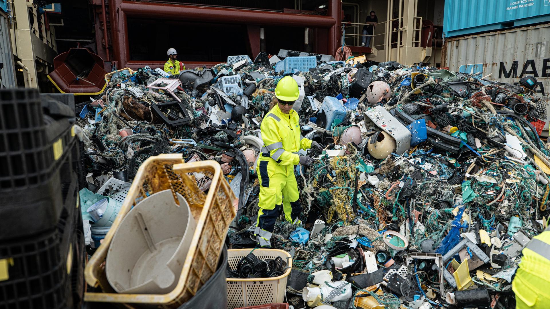 Eine Frau, im neon gelben Arbeitsanzug, steht am Deck eines Schiffes, inmitten eines Müllberges und sortiert Plastik.
