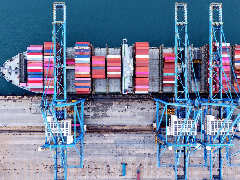 Viele bunte Container liegen an einem Hafen in Qingdao. Darüber stehen hellblaue Kräne. Das Bild wurde aus der Vogelperspektive aufgenommen.