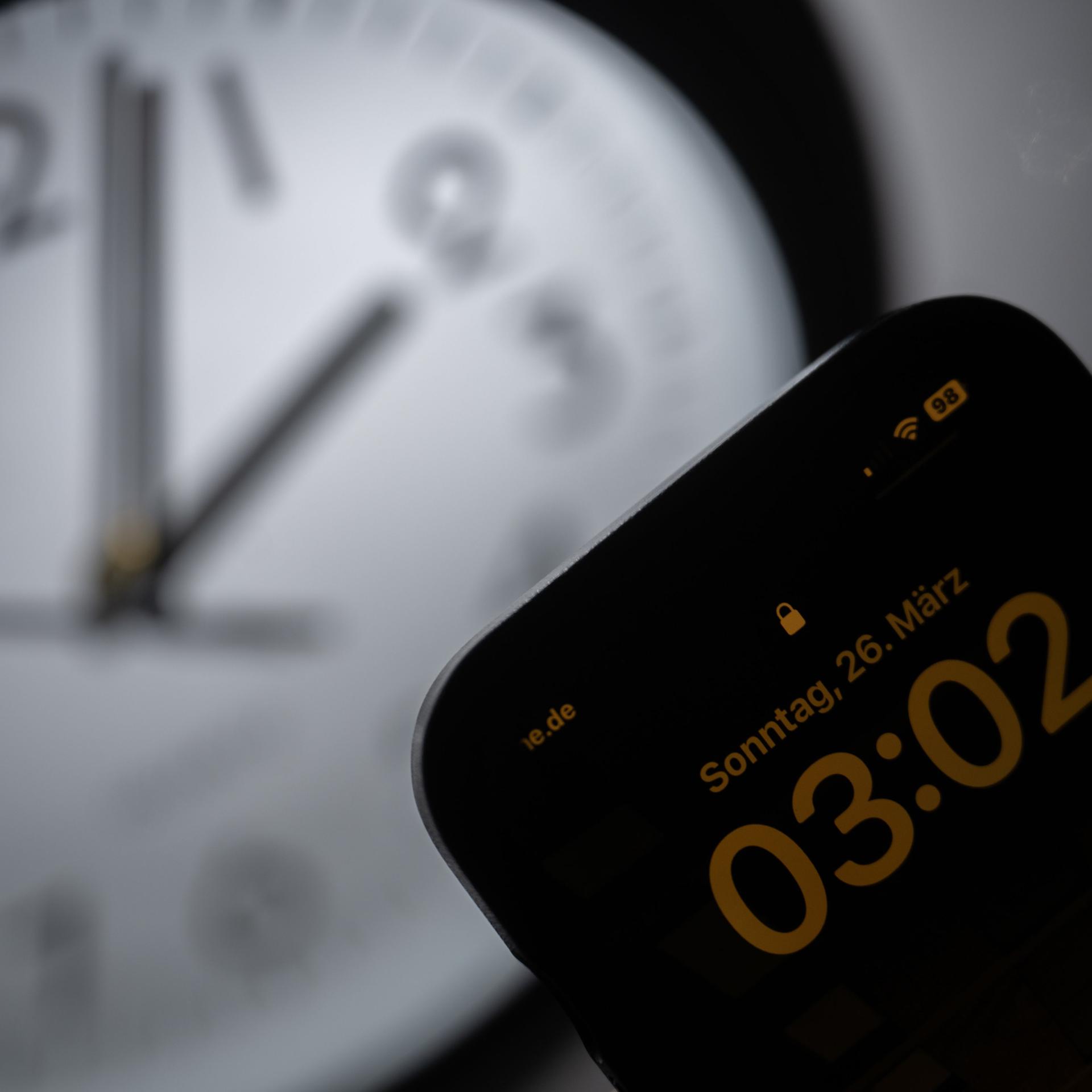 Ein Smartphone zeigt die Uhrzeit «03:03» an, während auf einer analogen Uhr noch 02:03 Uhr ist. 