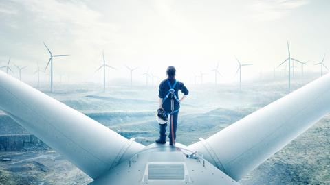 Eine Frau im Blaumann steht auf der Turbine eines Windrades und schaut aufs aufgewühlte Meer.