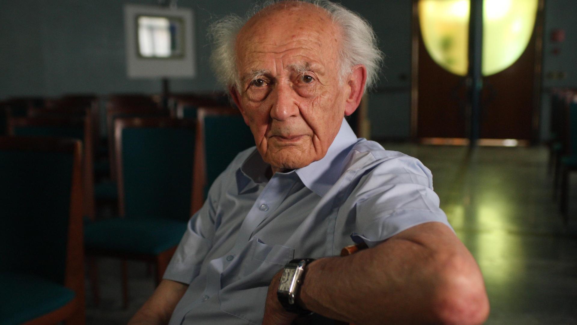 Der britisch-polnische Soziologe Zygmunt Bauman sitzt auf einem Stuhl und schaut seitlich gedreht direkt in die Kamera. 