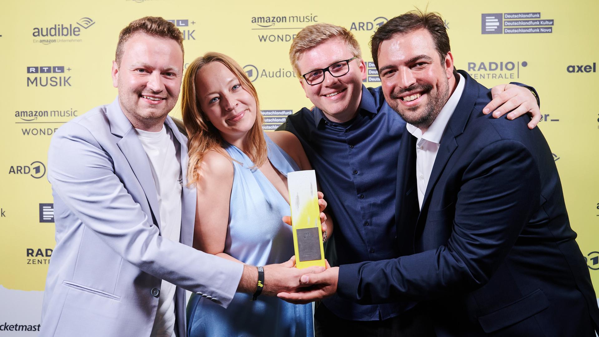 Marius Reichert, Laura Krzikalla, Till Krause und Constantin Pläcking stehen vor einer Fotowand mit dem Deutschen Podcast-Preis in den Händen.