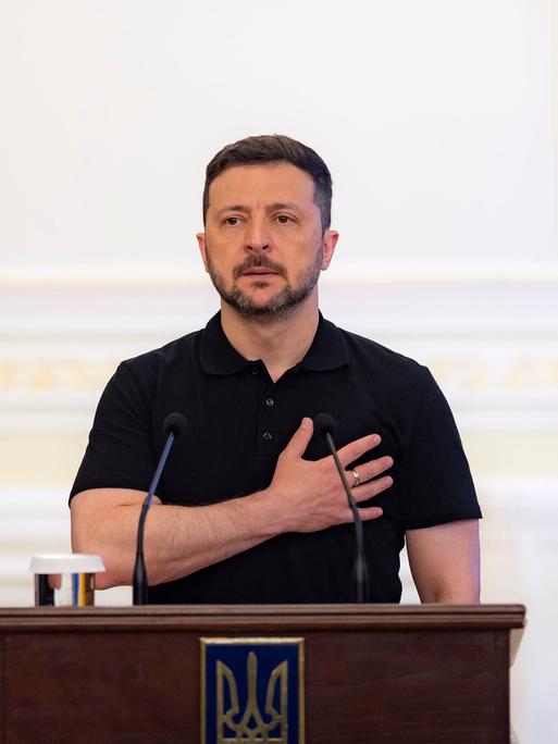 Der ukrainische Päsident Wolodymyr Selenskyj steht hinter einem Rednerpult und legt dabei die rechte Hand auf seinen Brustkorb.