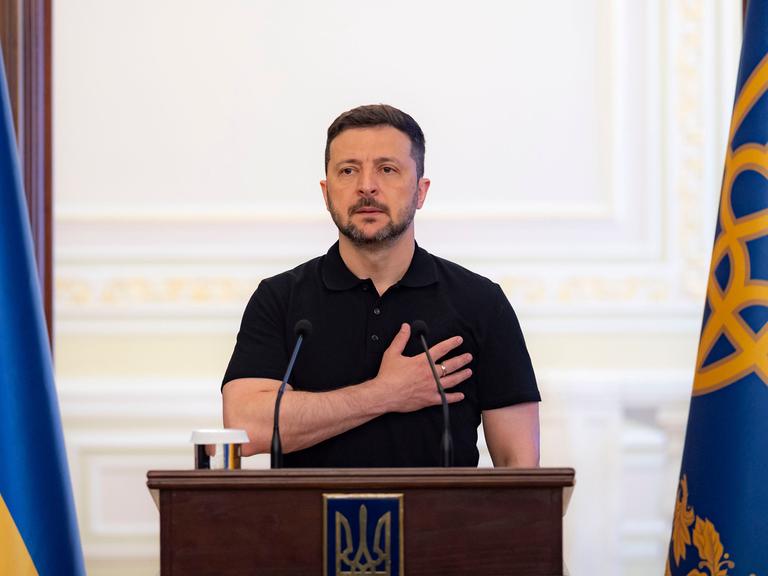 Der ukrainische Päsident Wolodymyr Selenskyj steht hinter einem Rednerpult und legt dabei die rechte Hand auf seinen Brustkorb.