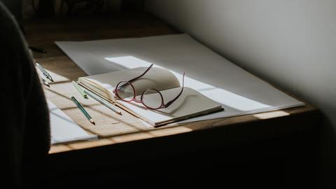 Eine Brille, die auf einem leeren Notizbuch liegt. Stifte umgeben das Buch, und Sonnenlicht fällt durch das Fenster und wirft Sonnenstrahlen auf den Schreibtisch. Eine Brille, die auf einem leeren Notizbuch liegt. Stifte umgeben das Buch, und Sonnenlicht fällt durch das Fenster und wirft Sonnenstrahlen auf den Schreibtisch.