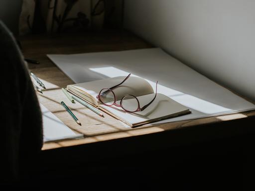 Eine Brille, die auf einem leeren Notizbuch liegt. Stifte umgeben das Buch, und Sonnenlicht fällt durch das Fenster und wirft Sonnenstrahlen auf den Schreibtisch.