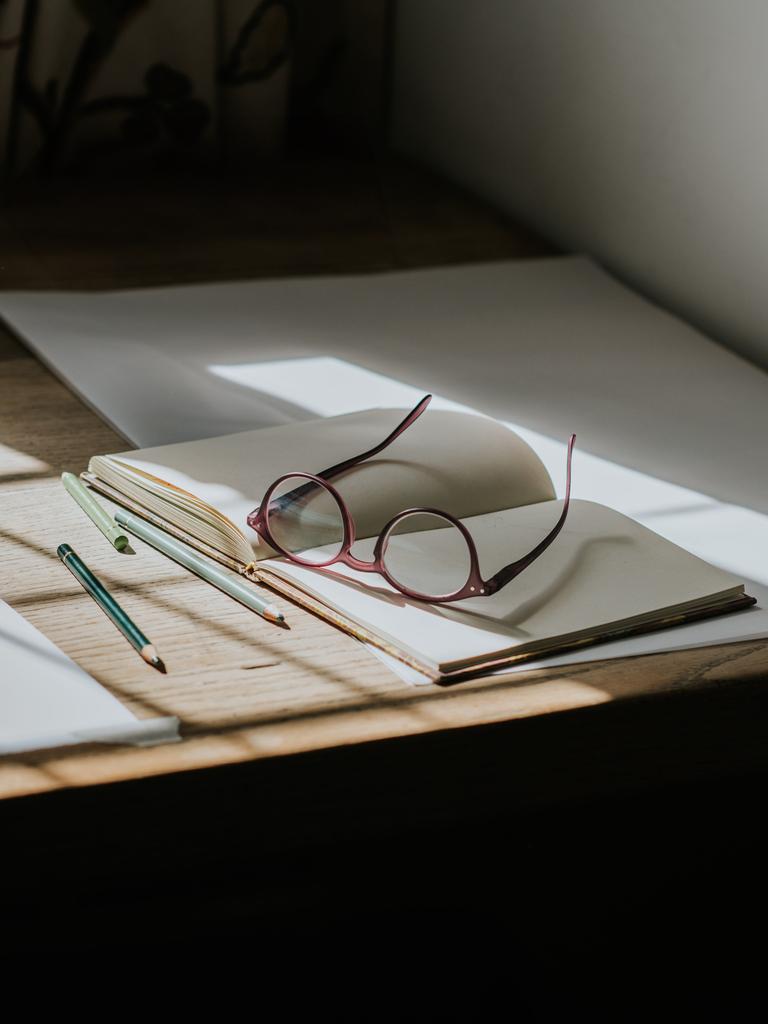 Eine Brille, die auf einem leeren Notizbuch liegt. Stifte umgeben das Buch, und Sonnenlicht fällt durch das Fenster und wirft Sonnenstrahlen auf den Schreibtisch.
